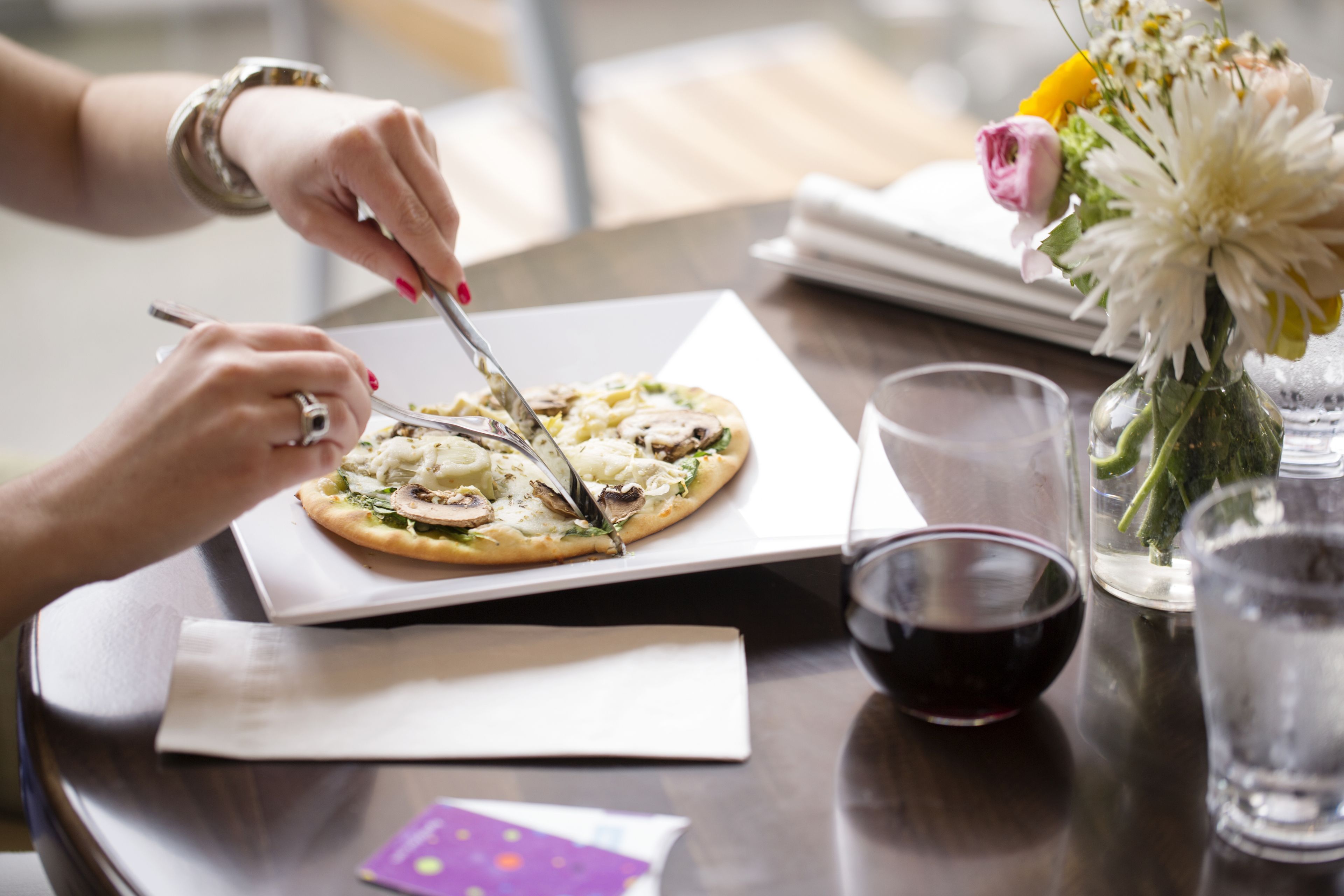 A person cutting a pizza on a white plate with a knife and fork. There is a glass of red wine and a vase with flowers on the table.