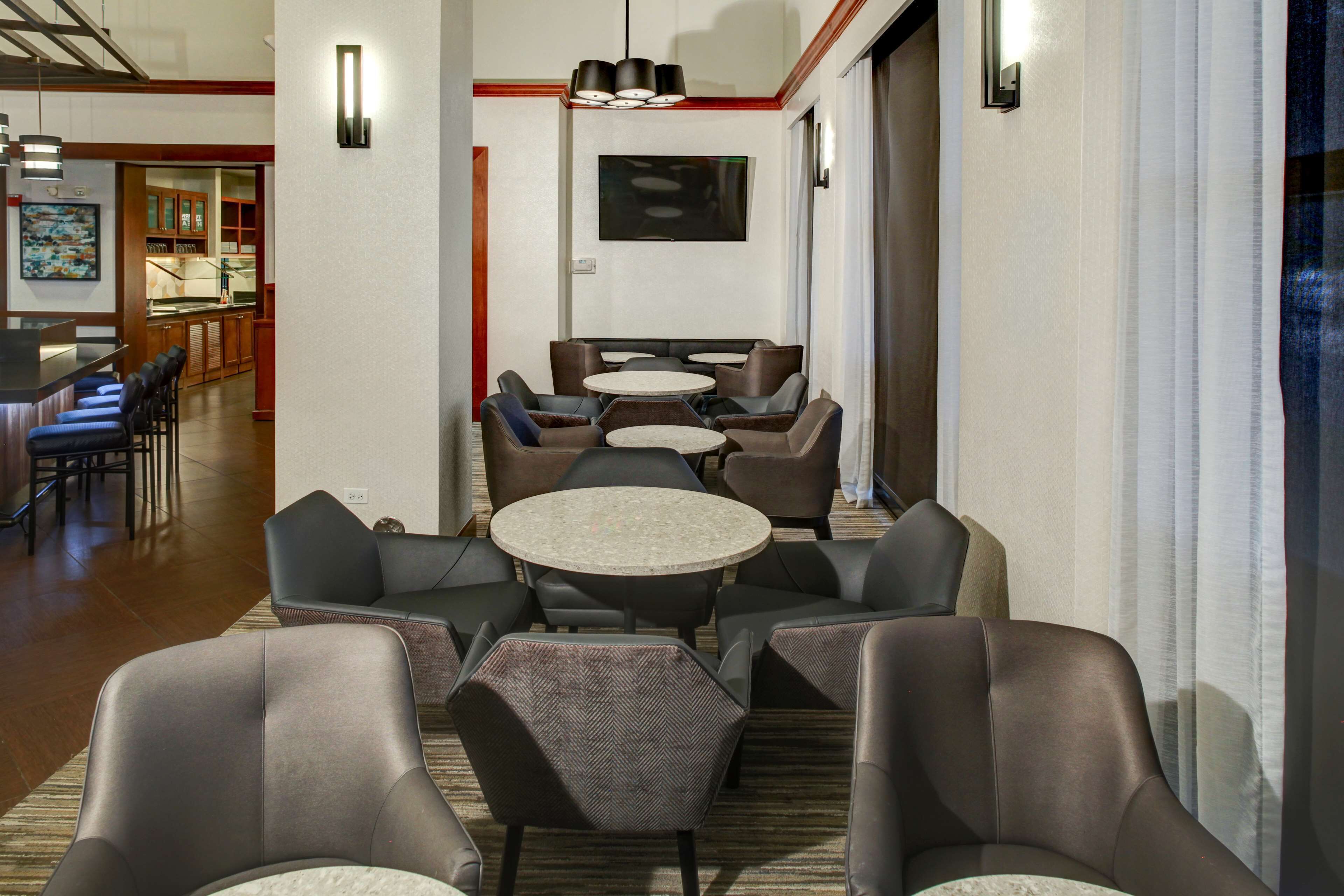 A hotel lobby with multiple seating areas, including round tables and chairs, a television mounted on the wall, and a bar area in the background.