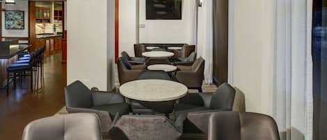 A hotel lobby with multiple seating areas, including round tables and chairs, a television mounted on the wall, and a bar area in the background.