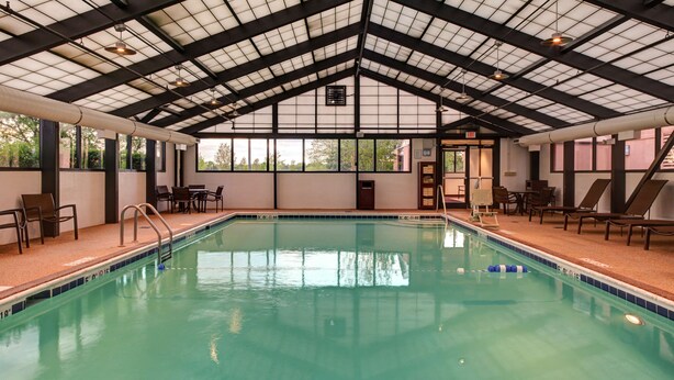 An indoor swimming pool with clear water, surrounded by lounge chairs and tables, under a high ceiling with a grid pattern. - Hyatt Place Itasca (Itasca)