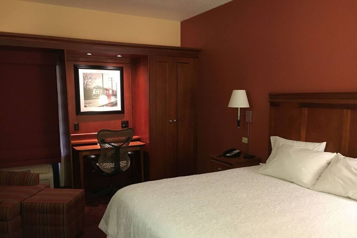 A hotel room with a large bed, a desk with a chair, a nightstand with a lamp, and a framed picture on the wall.