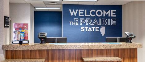Un mostrador de recepción con un letrero que dice “Welcome to the Prairie State”, una caja registradora y un quiosco de autoservicio.
