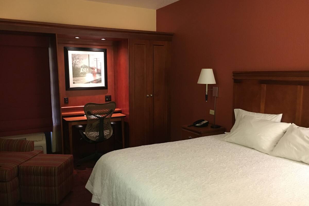 Een hotelkamer met een bed, een bureau met stoel, een nachtkastje met lamp en een lijst aan de muur.