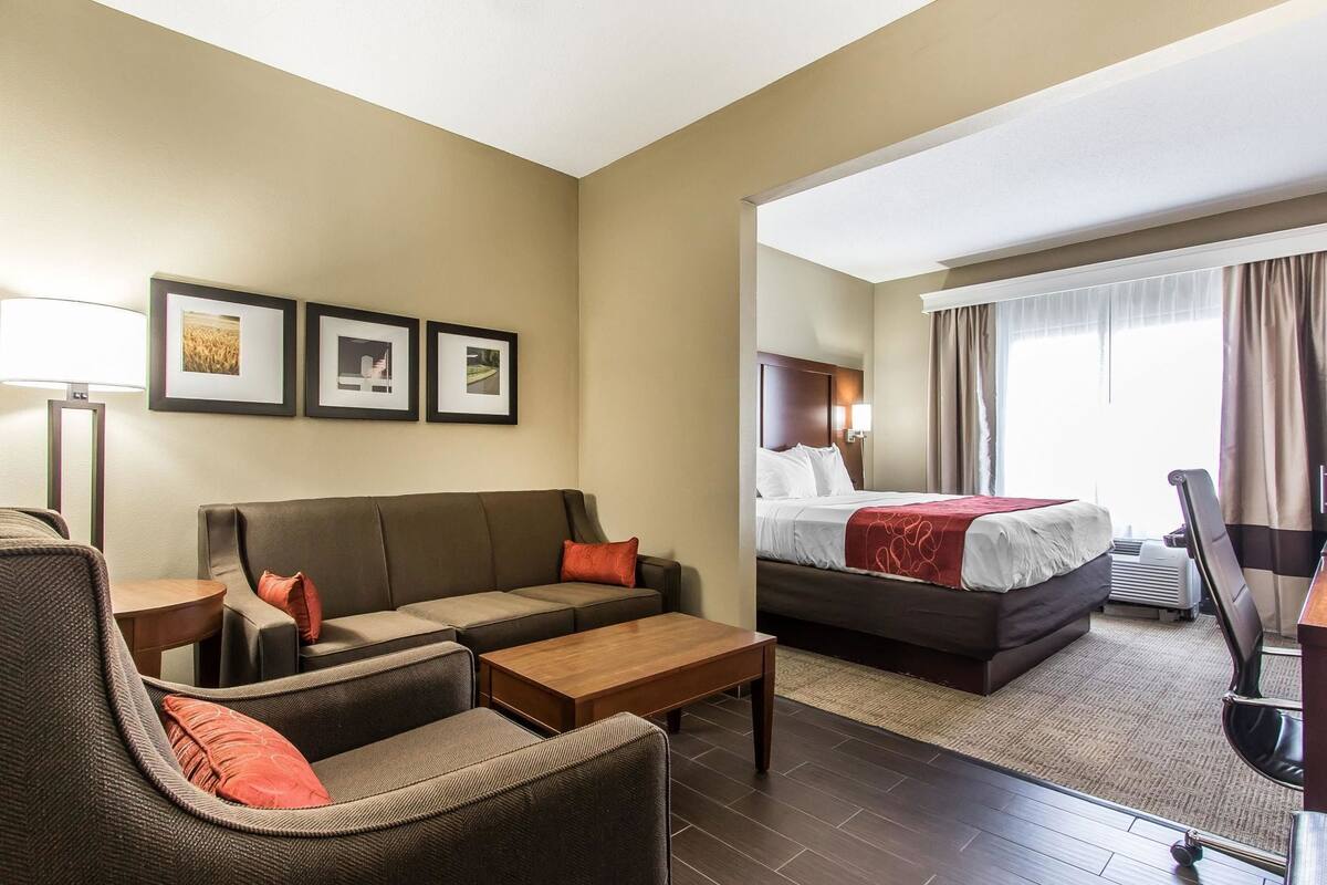 A hotel room with a sofa, a bed, a desk, and a chair. There are framed pictures on the wall and a window with curtains.
