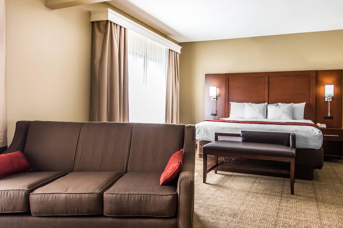 A hotel room with a brown sofa, a bed with white bedding, a wooden headboard, and a nightstand with a lamp.