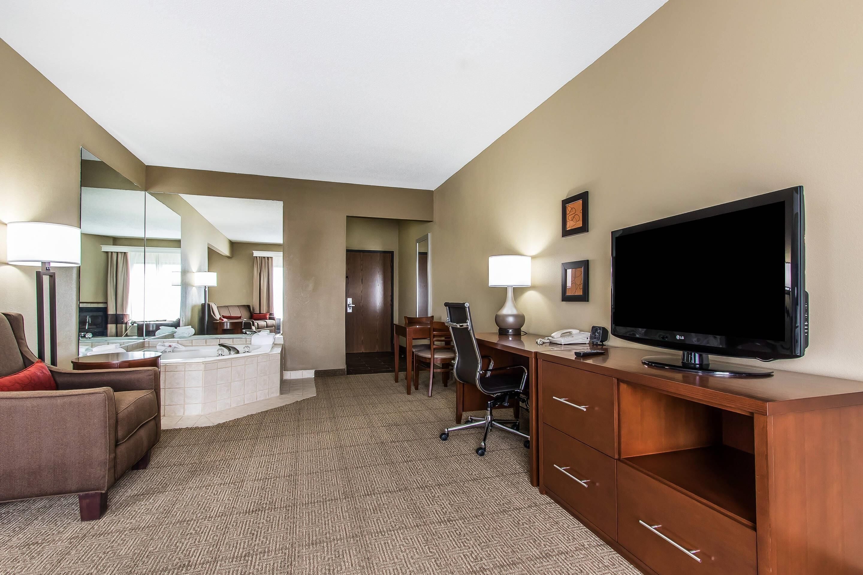 Habitación de hotel con una cama grande, un escritorio con televisor de pantalla plana, una silla, lámpara de escritorio, espejo y baño con lavamanos y espejo.