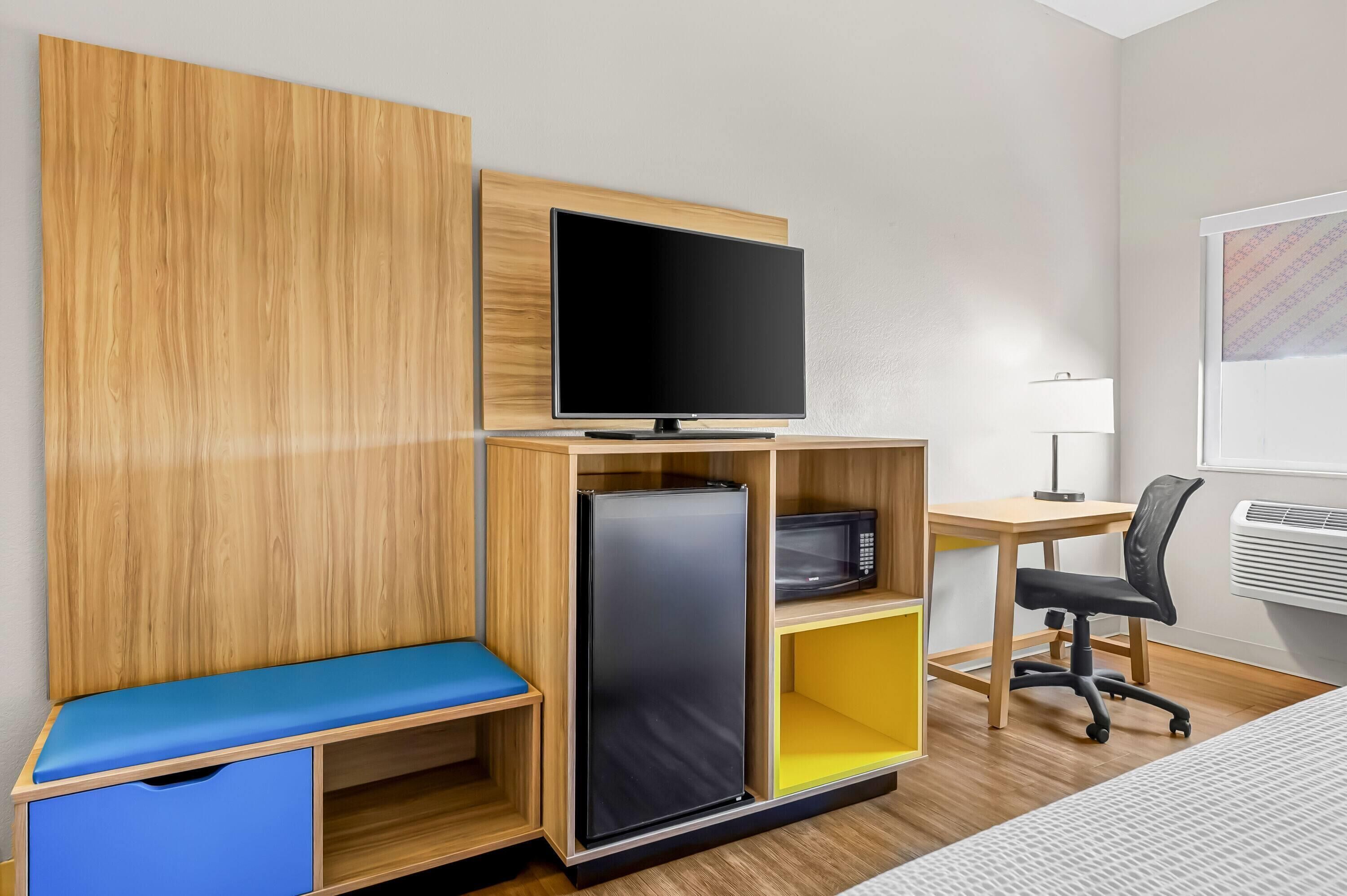 Une chambre de taille modeste comprenant un lit, un bureau avec un ordinateur, une télévision, un four à micro-ondes et un petit réfrigérateur.