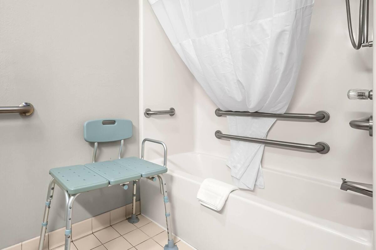 A bathroom with a shower, grab bars, a wheelchair-accessible bench, and a white curtain.