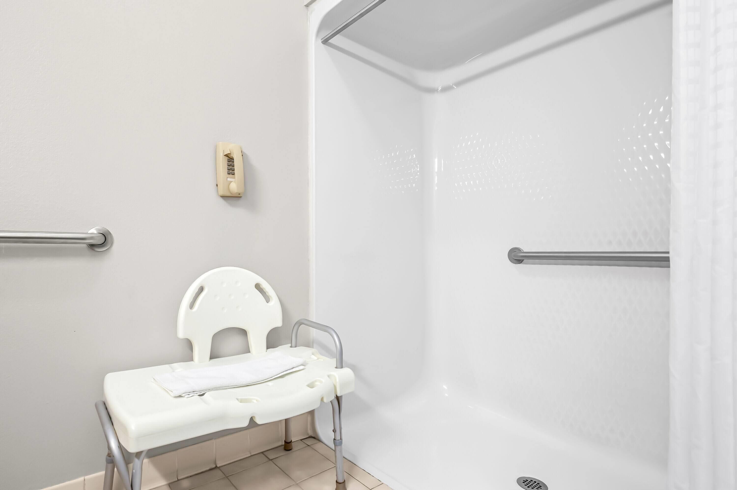 A shower area with a white shower seat, grab bars, and a handheld showerhead.