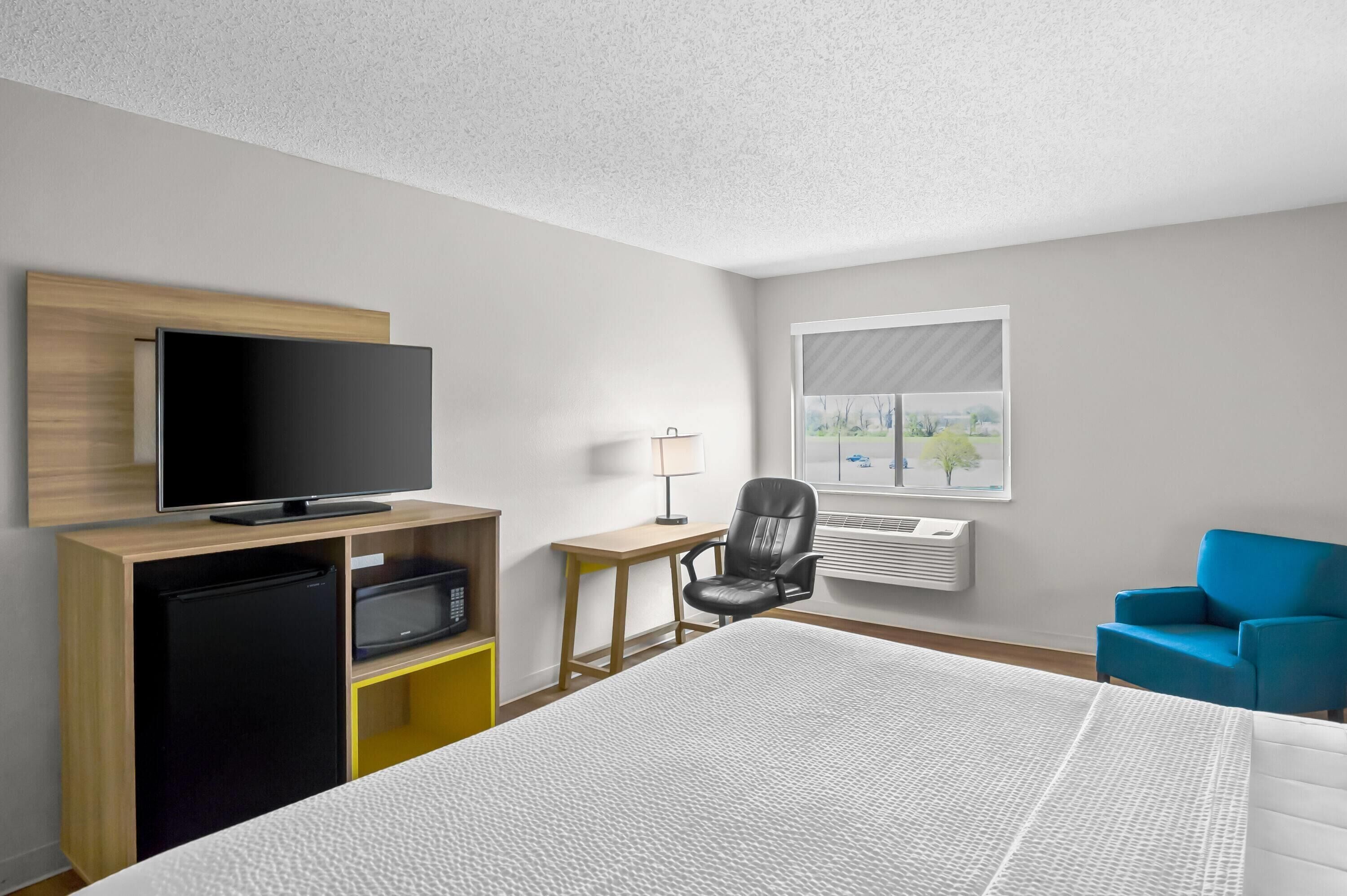 A hotel room with a bed, a TV on a wooden stand, a desk with a chair, a blue armchair, and a window with a view of trees.