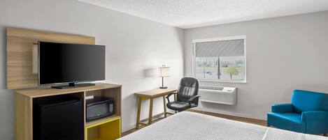 A hotel room with a bed, a TV on a wooden stand, a desk with a chair, a blue armchair, and a window with a view of trees.