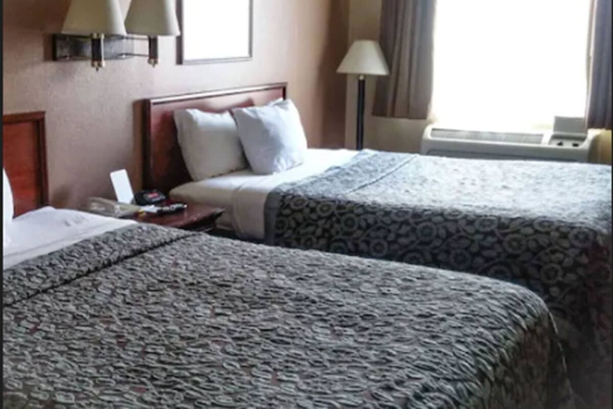 A hotel room with two beds, a nightstand, a lamp, and a window with curtains.