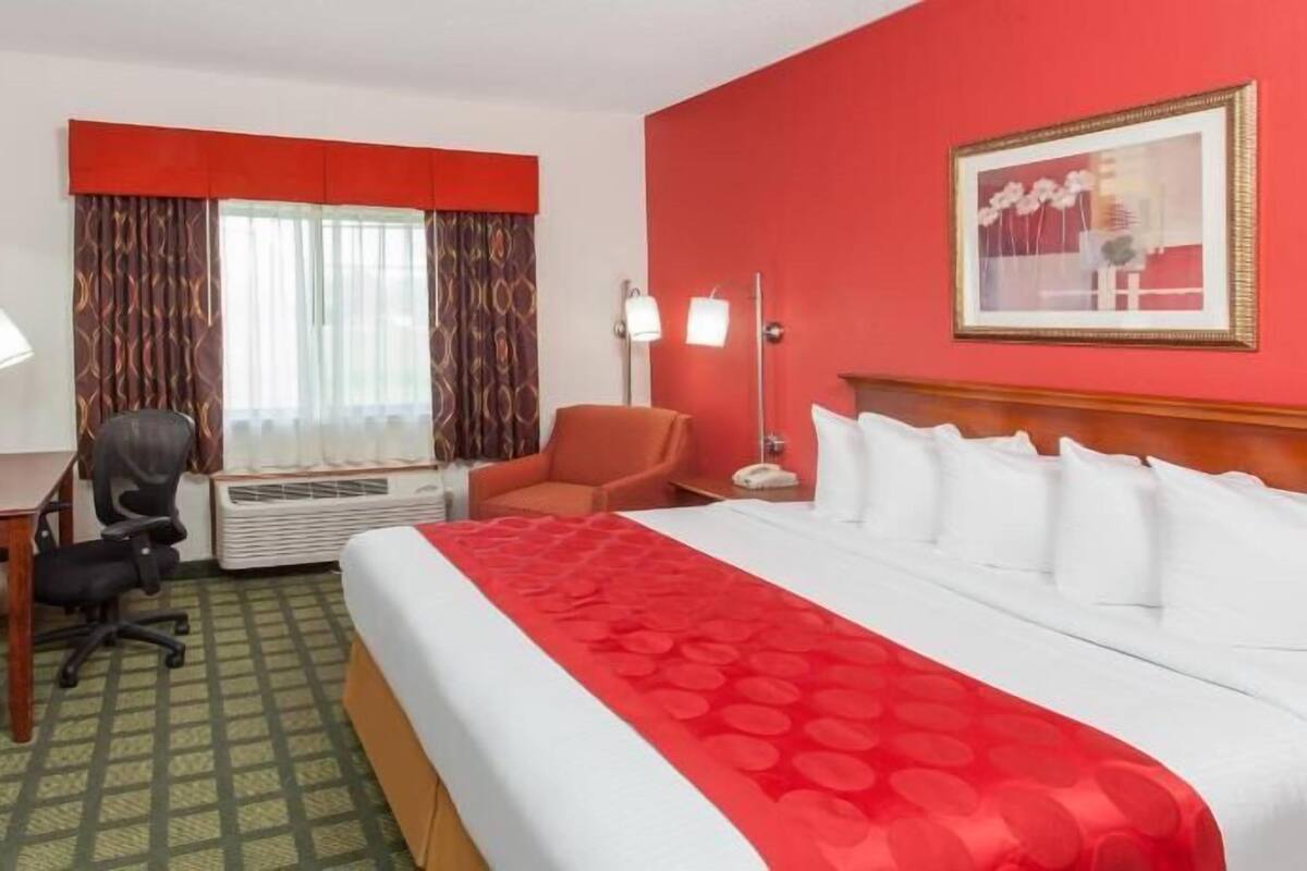 A hotel room with a bed, desk, chair, air conditioning unit, and a painting on the wall.