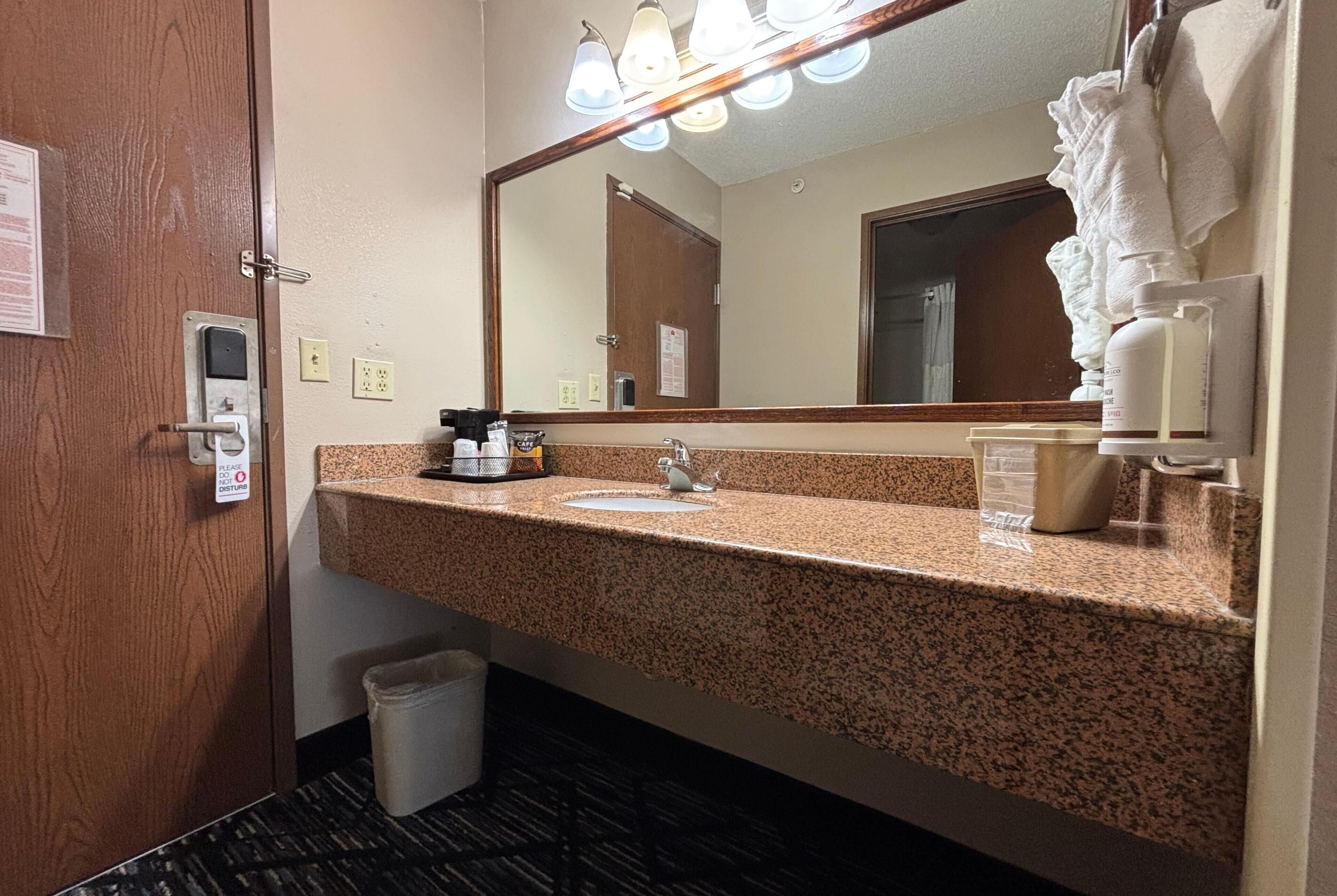 A hotel bathroom with a granite countertop, a sink, a mirror, a towel rack, and a trash bin.