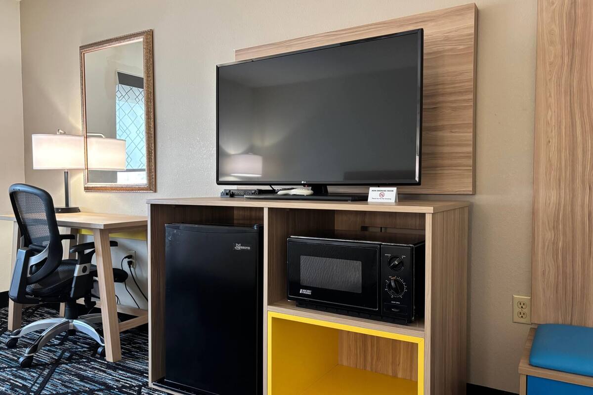 A hotel room with a desk, chair, television, microwave, and refrigerator.