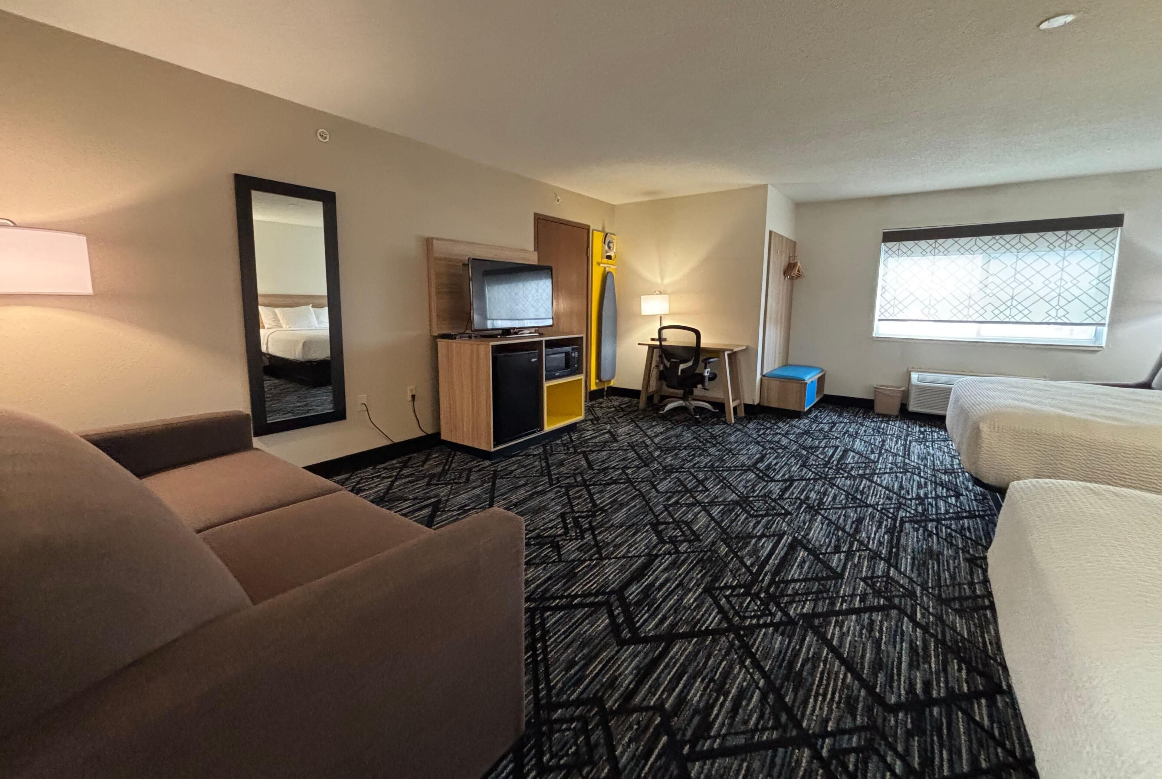 Une chambre d’hôtel avec un canapé, une télévision, un bureau avec une lampe, une fenêtre et un tapis à motifs.