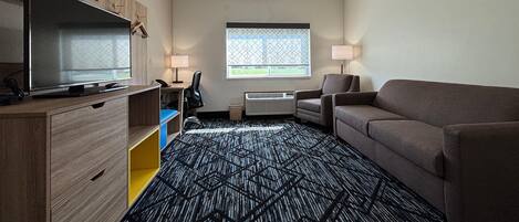 A hotel room with a ceiling fan, a flat-screen TV, a sofa, a desk with a chair, a window with blinds, and a patterned carpet.