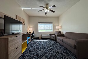 A hotel room with a ceiling fan, a flat-screen TV, a sofa, a desk with a chair, a window with blinds, and a patterned carpet. - Baymont by Wyndham Decatur IL North (Decatur)
