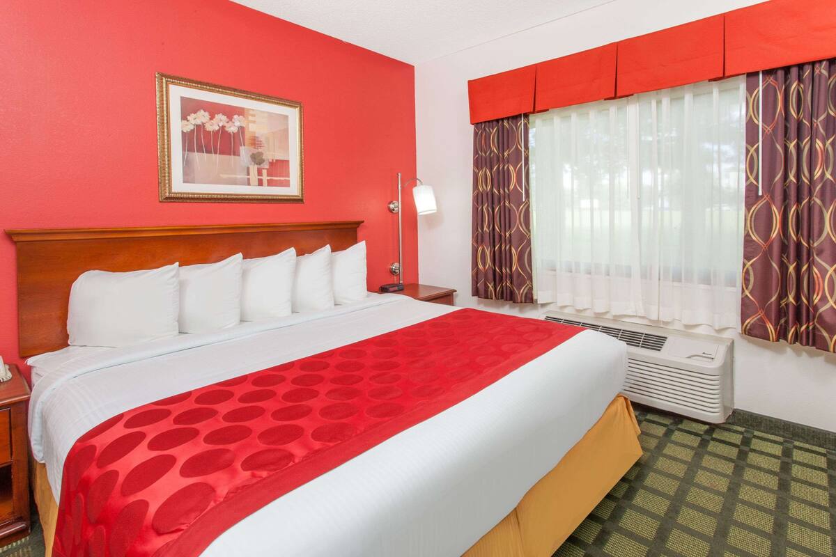 A hotel room with a large bed, a nightstand, a lamp, a window with curtains, and a wall-mounted air conditioner.
