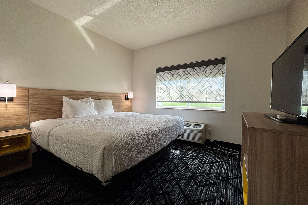 A hotel room with a bed, bedside lamp, window with view, air conditioning unit, and a TV on a wooden cabinet.