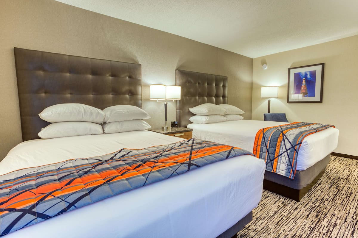 A hotel room with two beds, a headboard, bedside lamps, a framed picture on the wall, and a patterned rug.