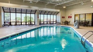An indoor swimming pool with clear glass walls, a tiled edge, and a ceiling with metal beams. There are lounge chairs and tables around the pool area.