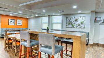 A modern bar area with a long wooden counter, cushioned bar stools, and a seating area with orange cushions.