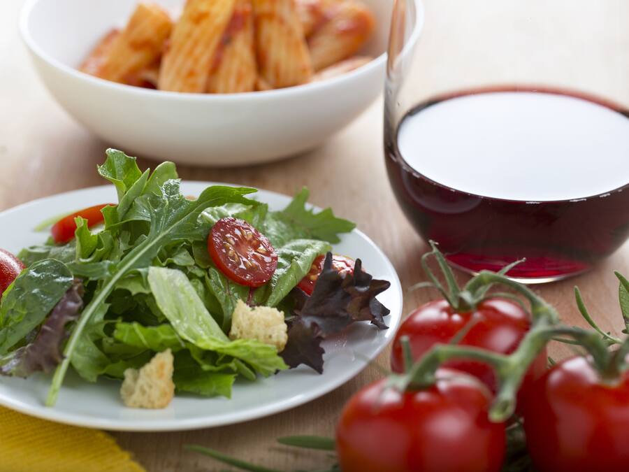 Un plato de ensalada de hojas verdes mezcladas con tomates cherry y croutons, un bol de pasta al horno y una copa de vino tinto sobre una mesa de madera.