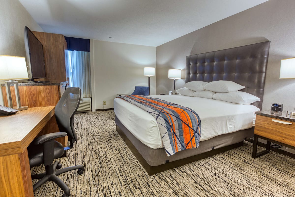 A hotel room with a bed, a desk, a chair, a lamp, and a clock.