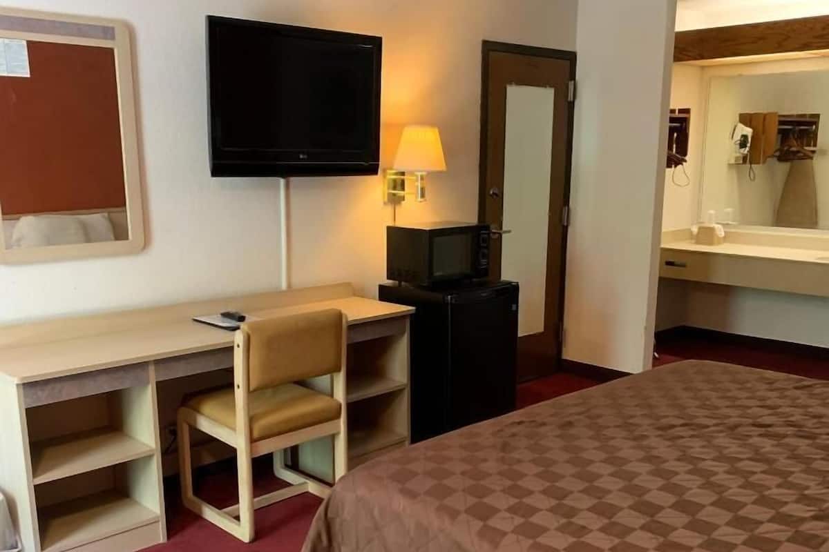 Una habitación de hotel con cama, escritorio con silla, televisor, lámpara y espejo.