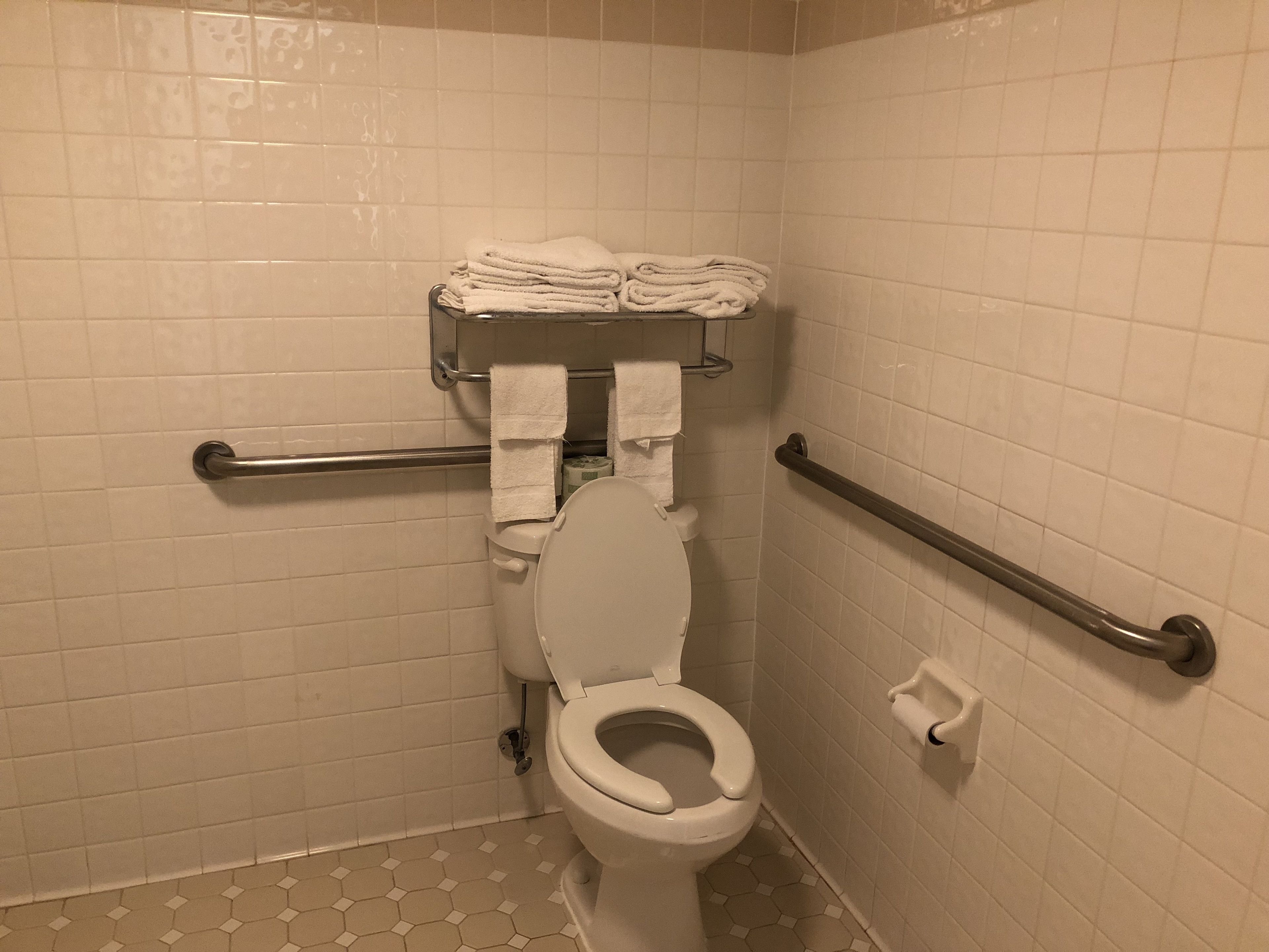 A bathroom with white tiled walls, a toilet with a raised seat, a grab bar, and a towel rack with folded towels.