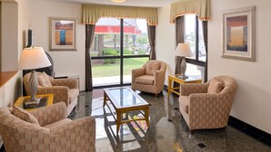 A hotel room with a seating area featuring four armchairs, a glass-top coffee table, and two lamps. Large windows provide a view of the outside.