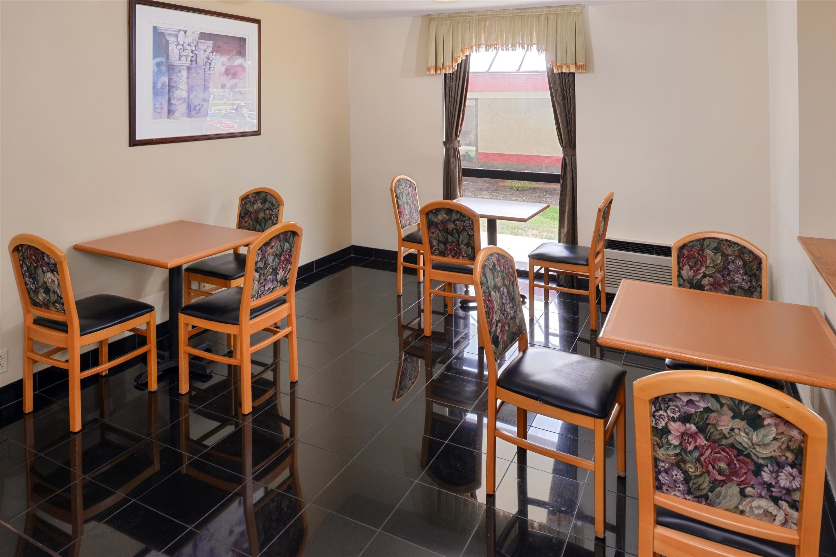 A dining area with wooden tables and chairs, a framed picture on the wall, and a window with curtains.