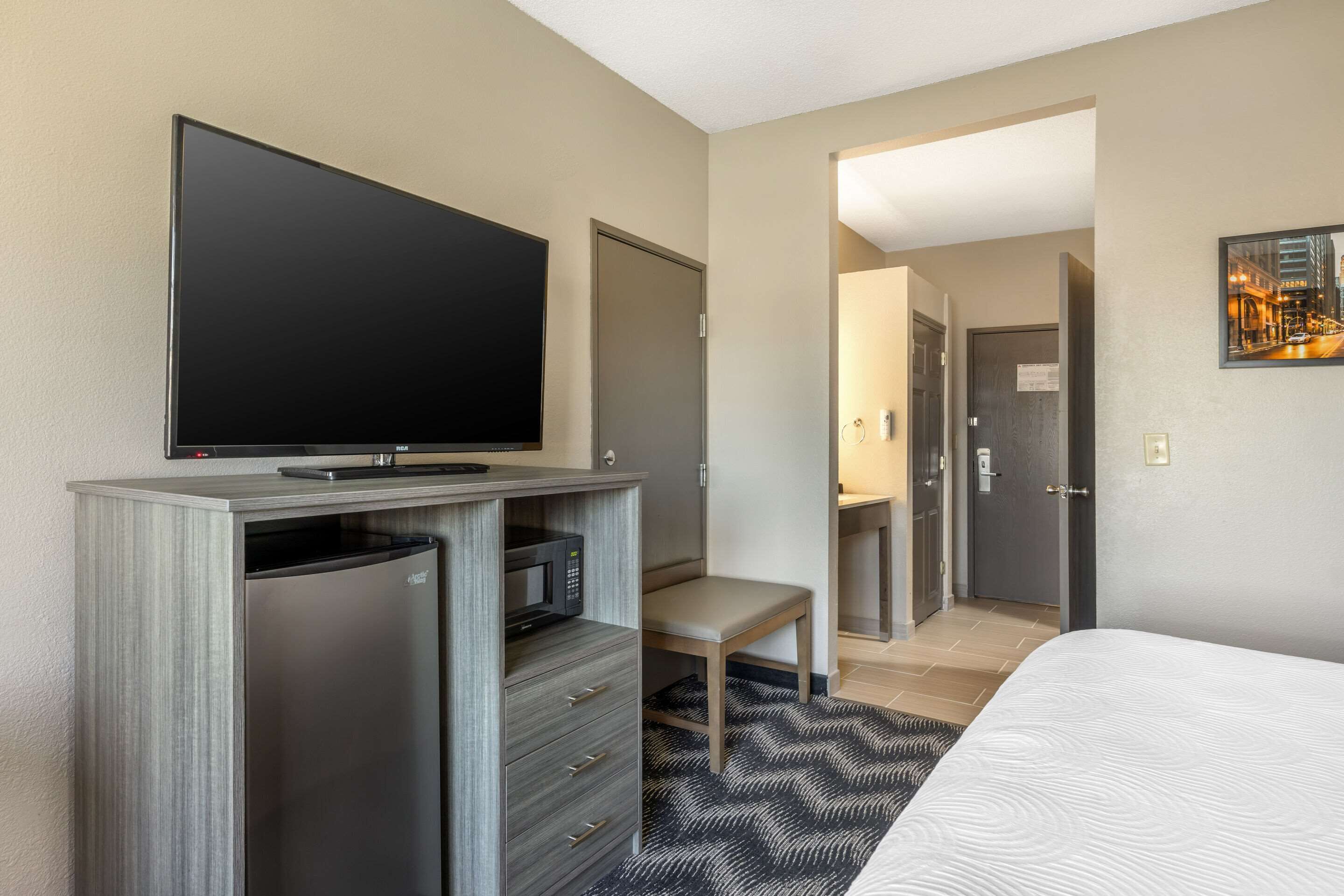 A hotel room with a flat-screen TV, a mini-fridge, a microwave, a bed, a desk, and a chair.