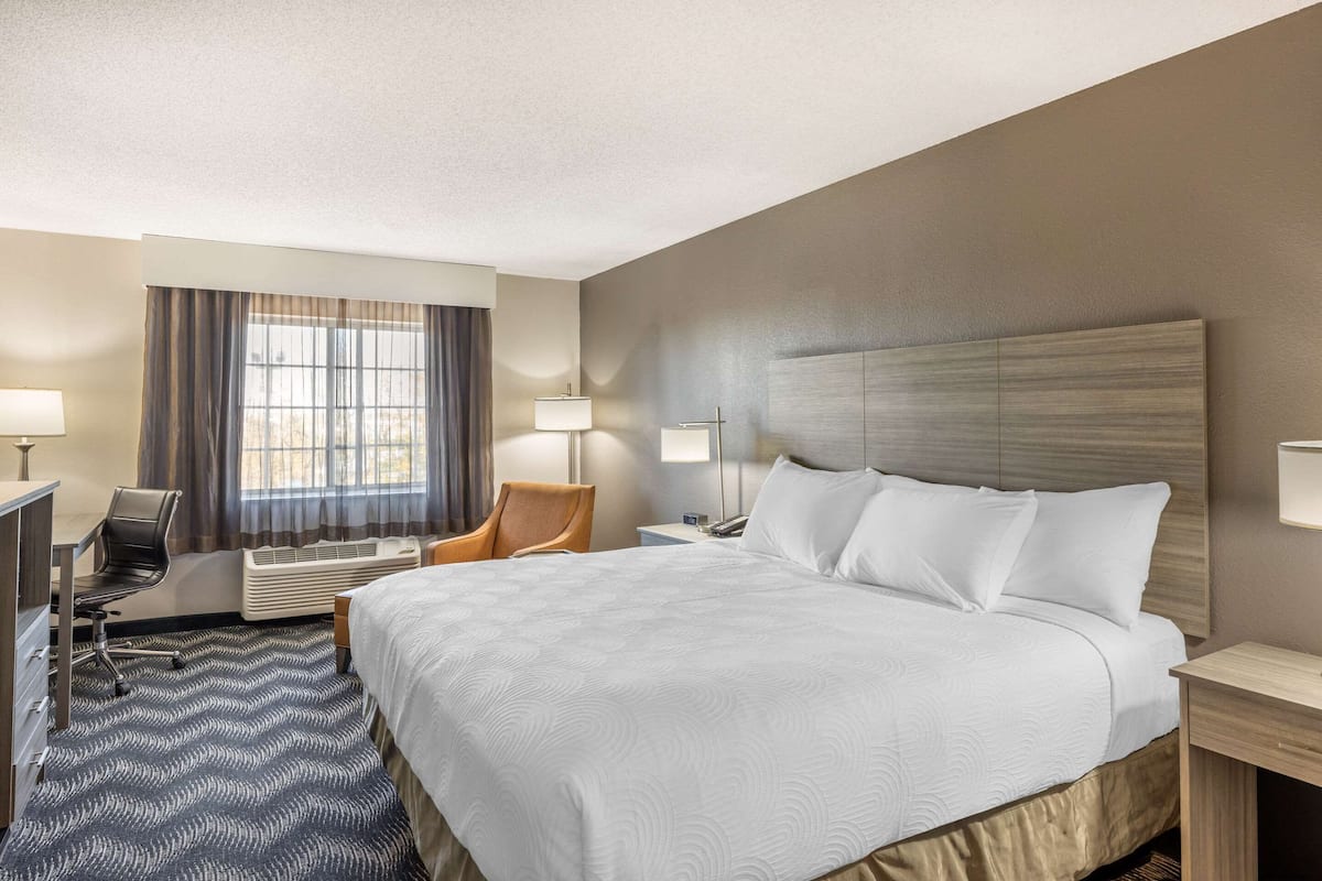 A hotel room with a large bed, a desk with a chair, a window with curtains, and a wall-mounted lamp.