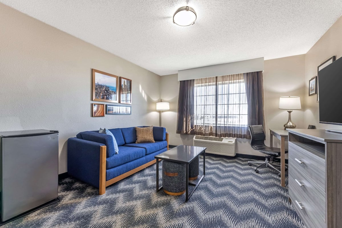 A compact hotel room with a blue sofa, a small table, a TV, a desk, and a refrigerator.
