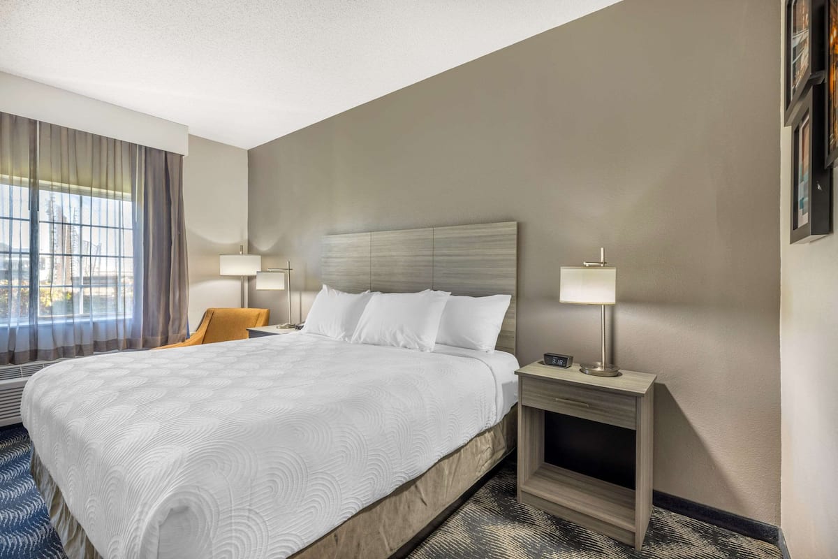 A hotel room with a large bed, two bedside tables with lamps, a chair, and a view of the city through large windows.