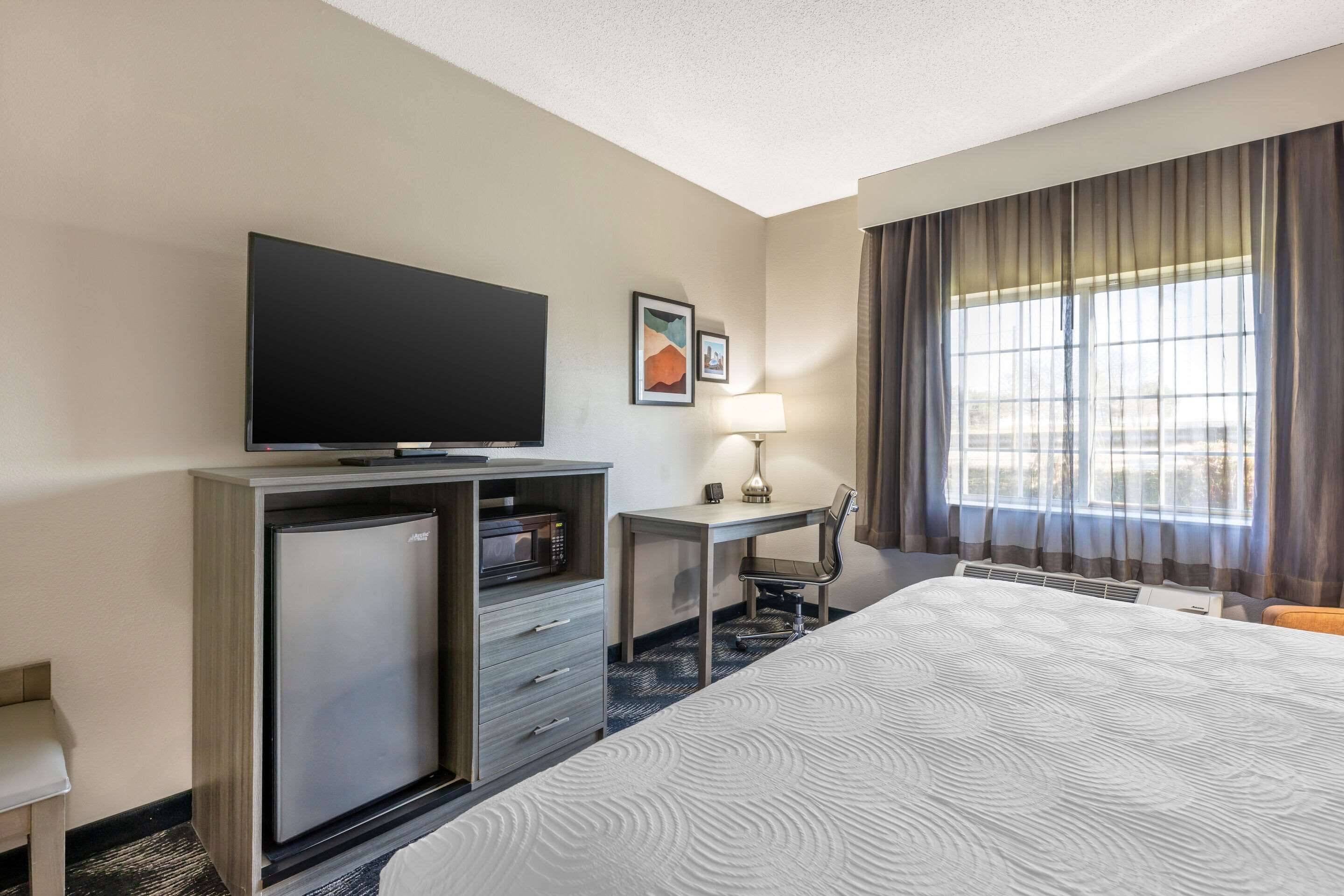 A hotel room with a bed, a desk, a television, a refrigerator, and a window with curtains.