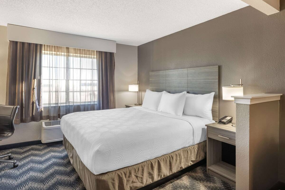 A hotel room with a large bed, a desk, a chair, a lamp, and a window with curtains.