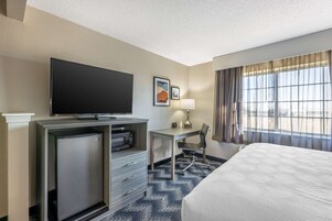 Habitación de hotel con cama, escritorio con silla, televisor, refrigerador pequeño y ventana con cortinas.