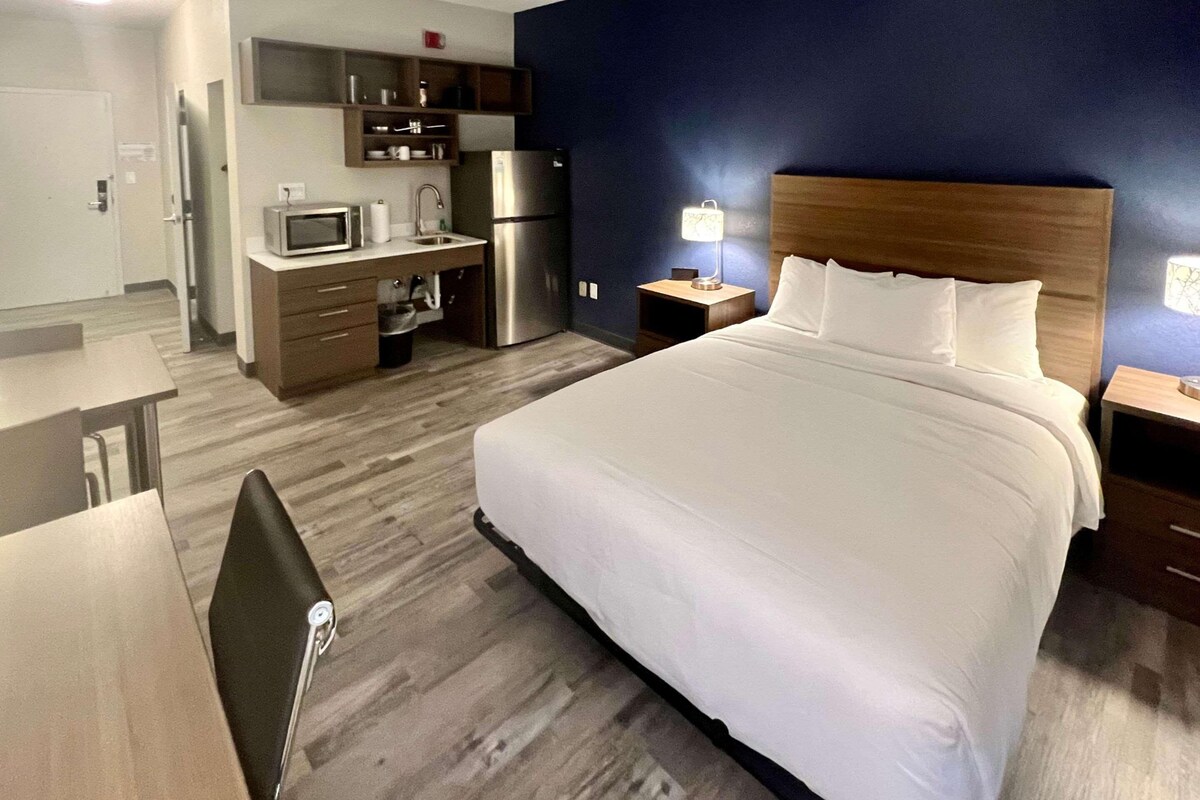 Habitación de hotel con cama, escritorio, microondas, refrigerador, mesita de noche y lámpara.