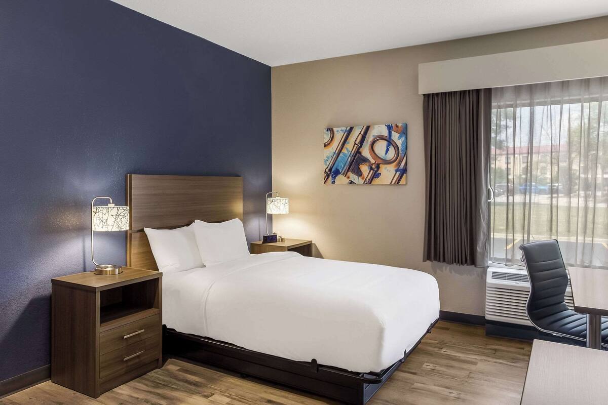 Une chambre d’hôtel avec un lit, des tables de chevet, une lampe, un tableau, une chaise et une fenêtre avec des rideaux.