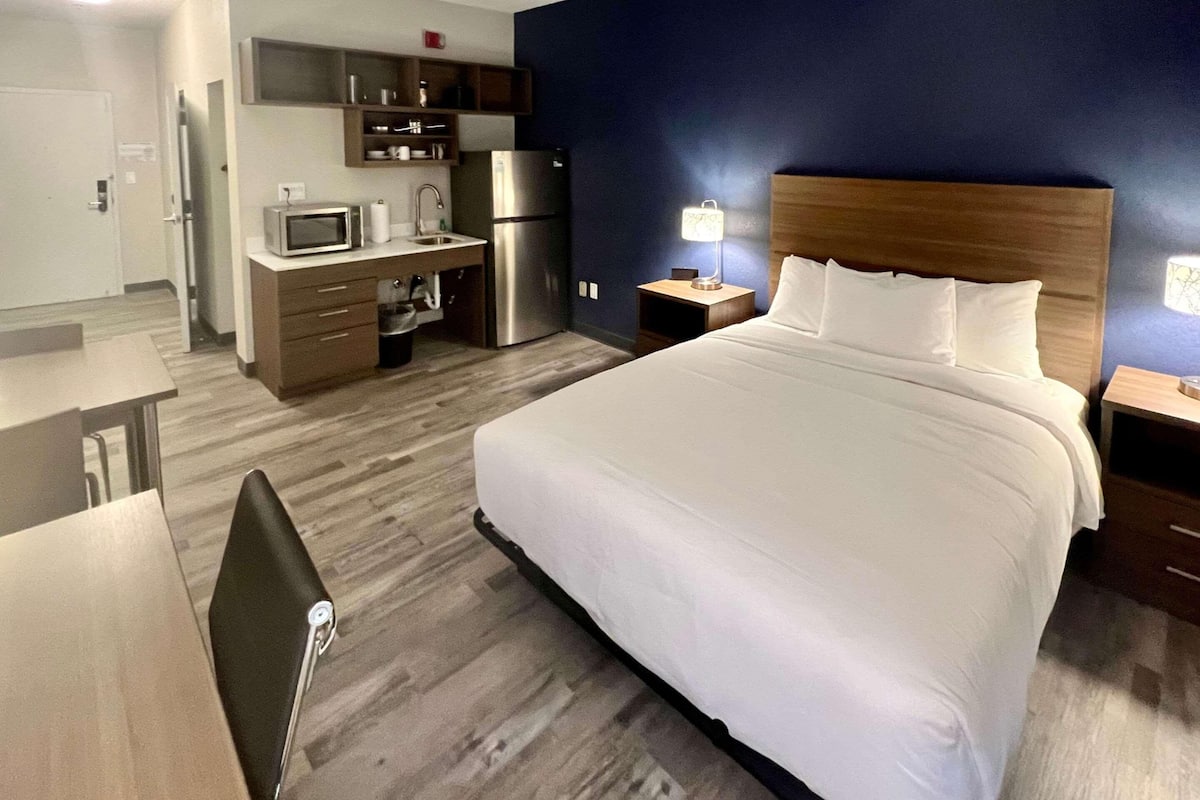 Habitación de hotel con cama, escritorio, microondas, refrigerador, mesita de noche y lámpara.