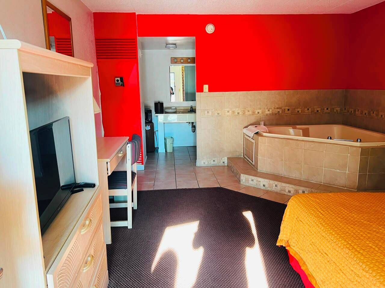 Habitación de hotel con televisión, escritorio, silla, cama y baño con bañera, y una pared roja.