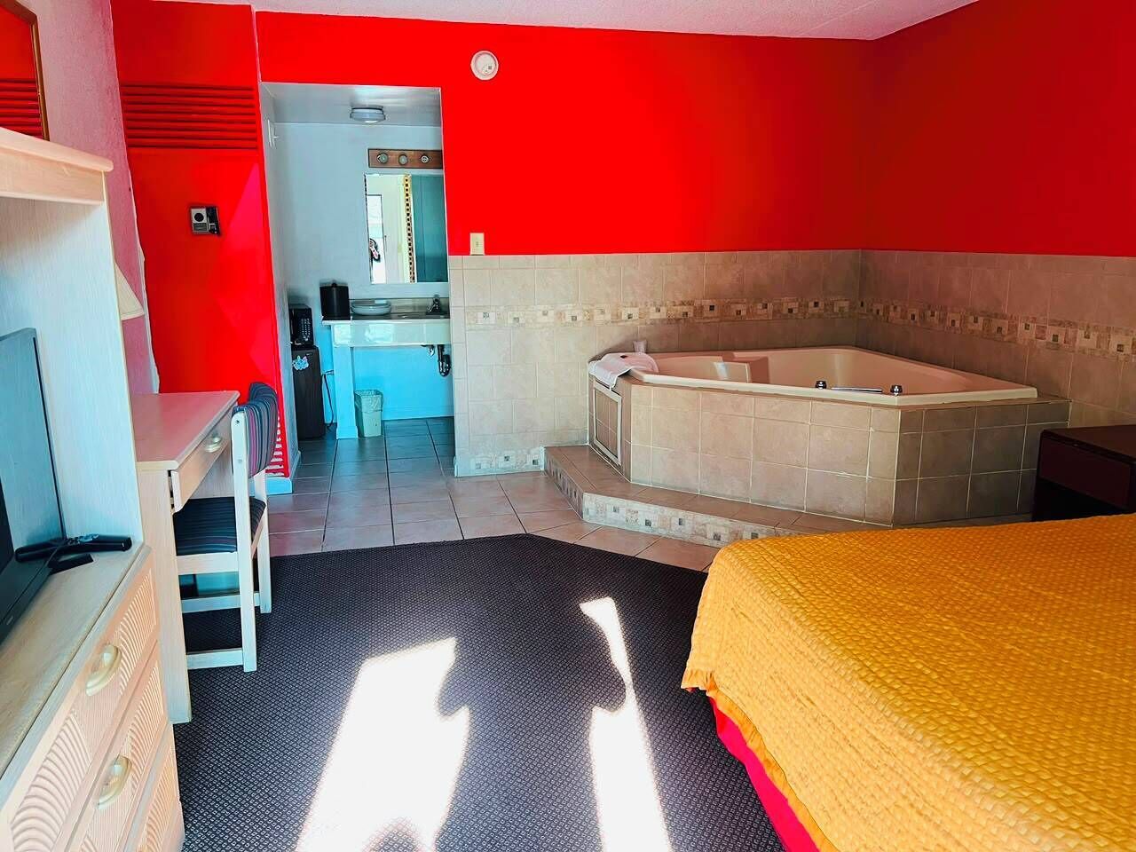 A hotel room with a bed, a desk, a chair, a small kitchenette, and a bathtub.