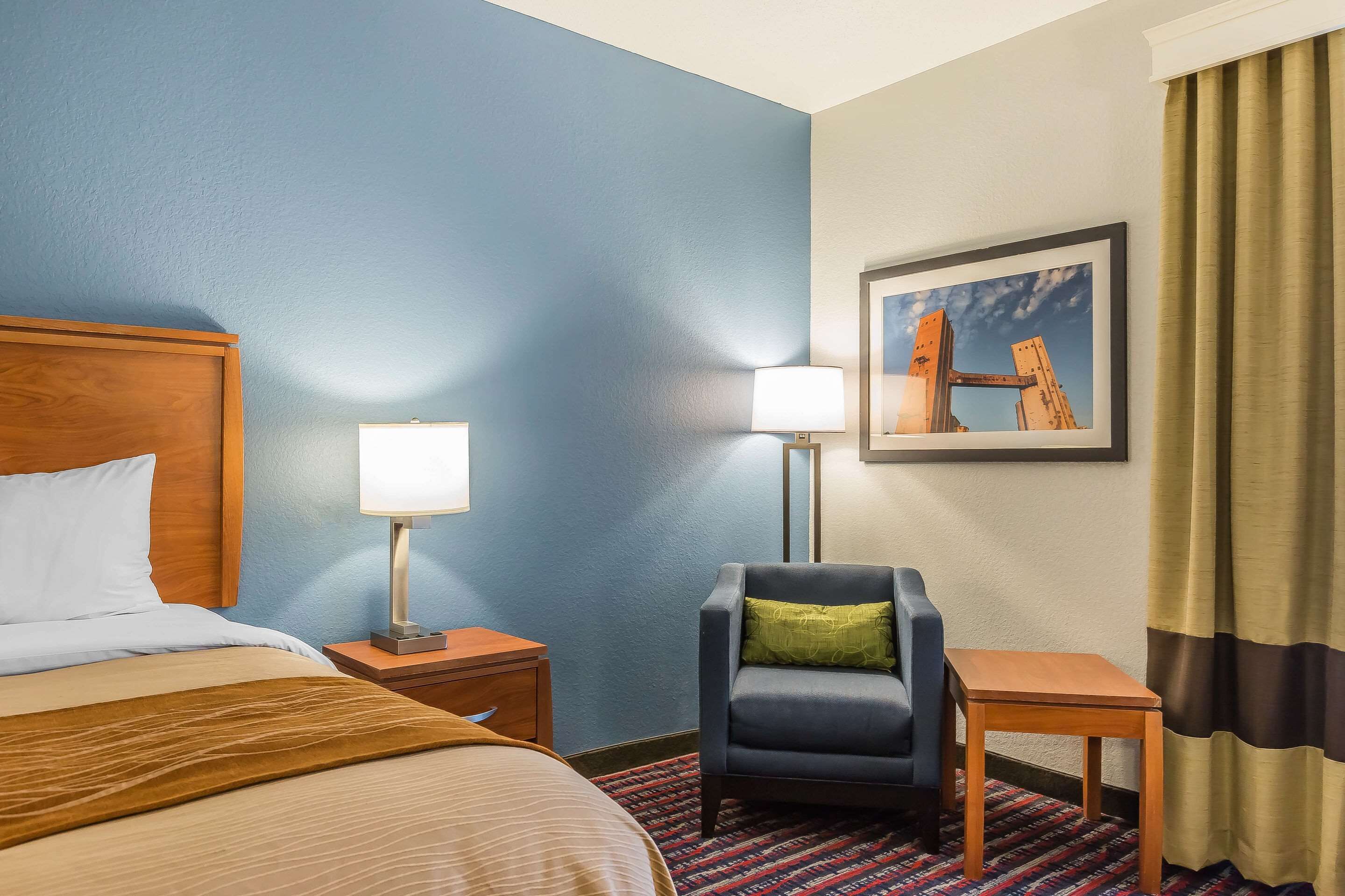 Quarto de hotel com cama, duas mesas de cabeceira com abajures, uma cadeira, uma mesinha e um quadro na parede, além de um tapete estampado.