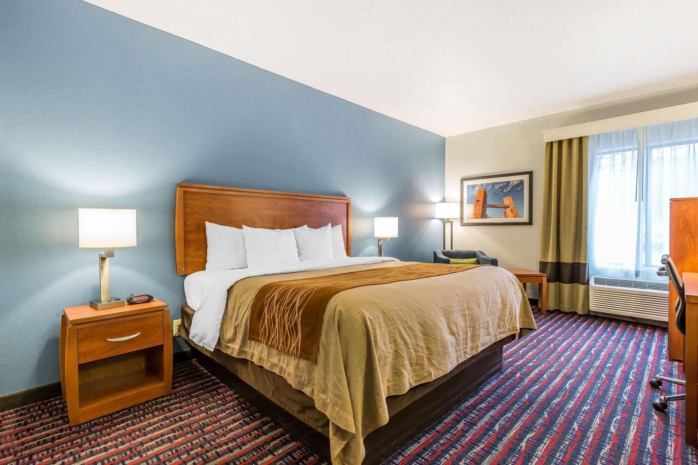 Habitación de hotel con cama, mesitas de noche, escritorio, silla, ventana con cortinas y un cuadro en la pared.