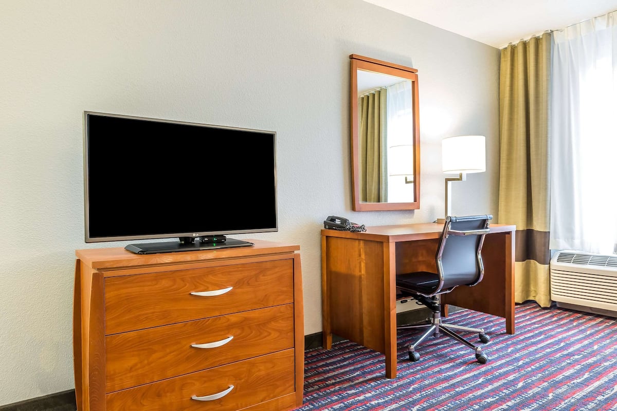 A hotel room with a wooden desk, a chair, a television, a mirror, and a lamp.