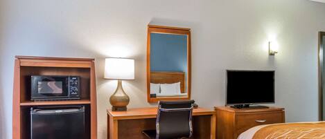 Een hotelkamer met een bed, een bureau met een computer, een koelkast, een magnetron, een televisie en een spiegel.