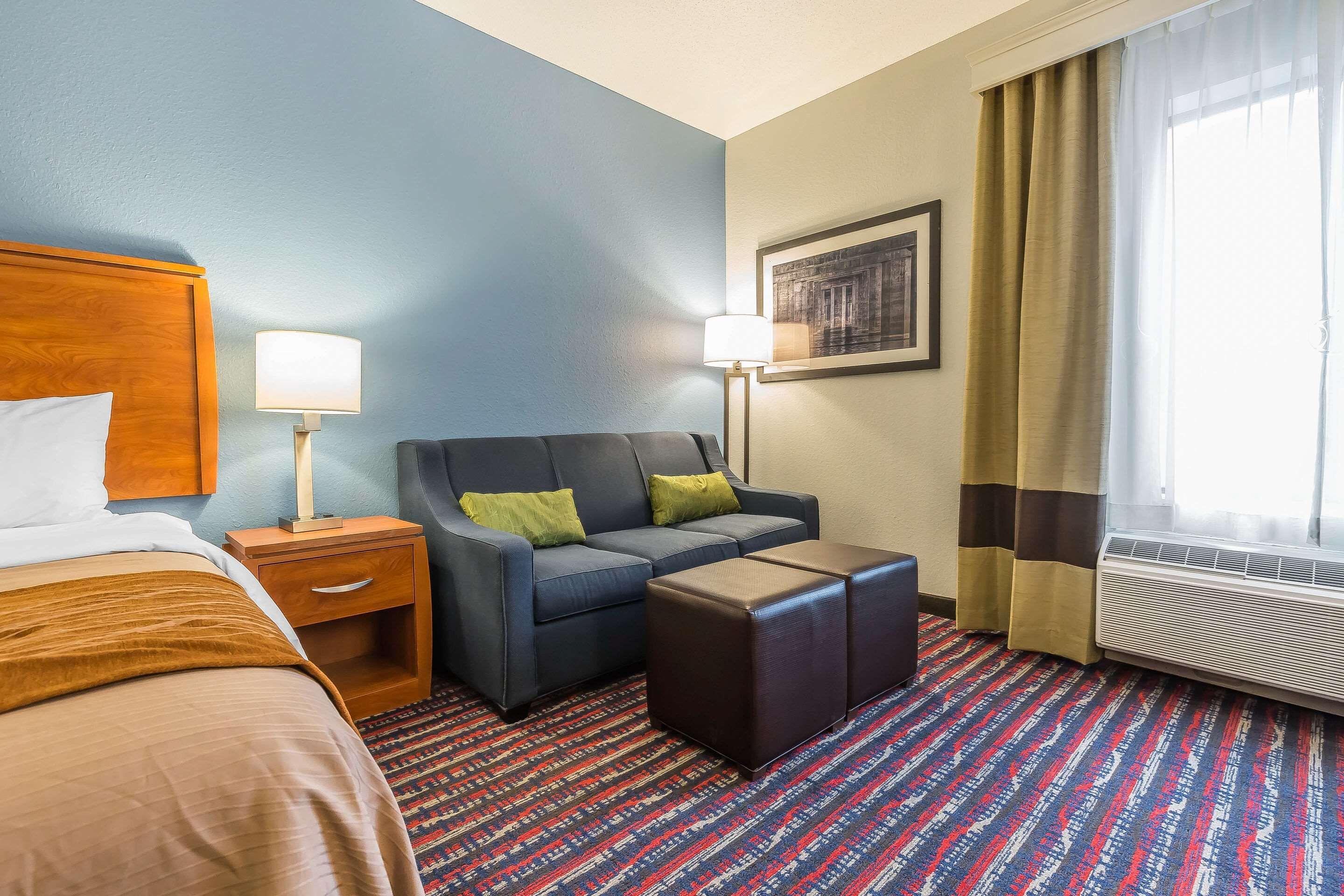 Habitación de hotel con cama, sofá, mesita, mesita de noche, lámpara, aire acondicionado y ventana con cortinas.